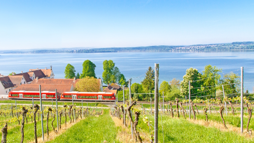 Blick auf den Bodensee vom Kloster Birnau aus. Im Vordergrund befindet sich ein Weinberg. Am Fuß des Hügels passiert eine Regionalbahn einige Häuser. Blick auf den Bodensee vom Kloster Birnau aus. Im Vordergrund befindet sich ein Weinberg. Am Fuß des Hügels passiert eine Regionalbahn einige Häuser.